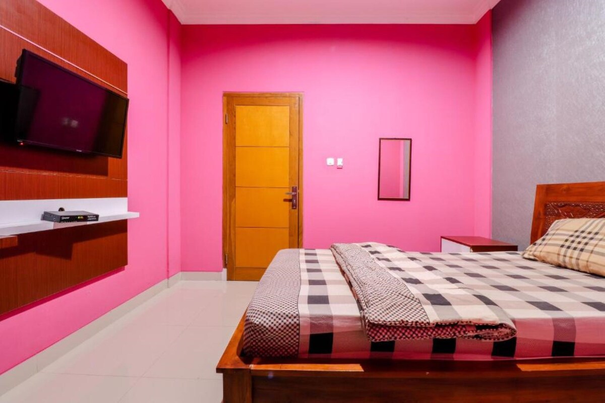 A bedroom with a pink wall, a wooden bed frame, a checkered bedspread, a television mounted on a wooden shelf, and a door with a wooden frame.