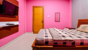 A bedroom with a pink wall, a wooden bed frame, a checkered bedspread, a television mounted on the wall, and a wooden door. - Khatarina Female Residence - Female Only - khusus wanita (Depok)