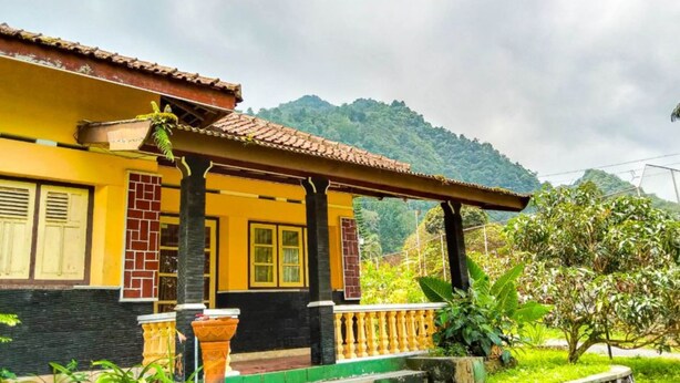 Um edifício amarelo com varanda, pilares de tijolo e escadaria verde que leva à entrada.
