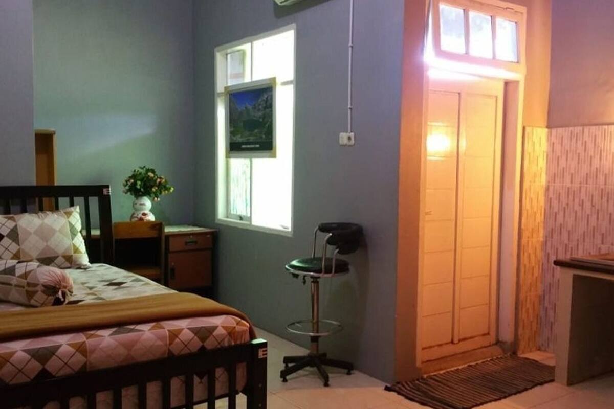 Un dormitorio con cama, mesita de noche, silla, ventana y puerta que da a otra habitación.