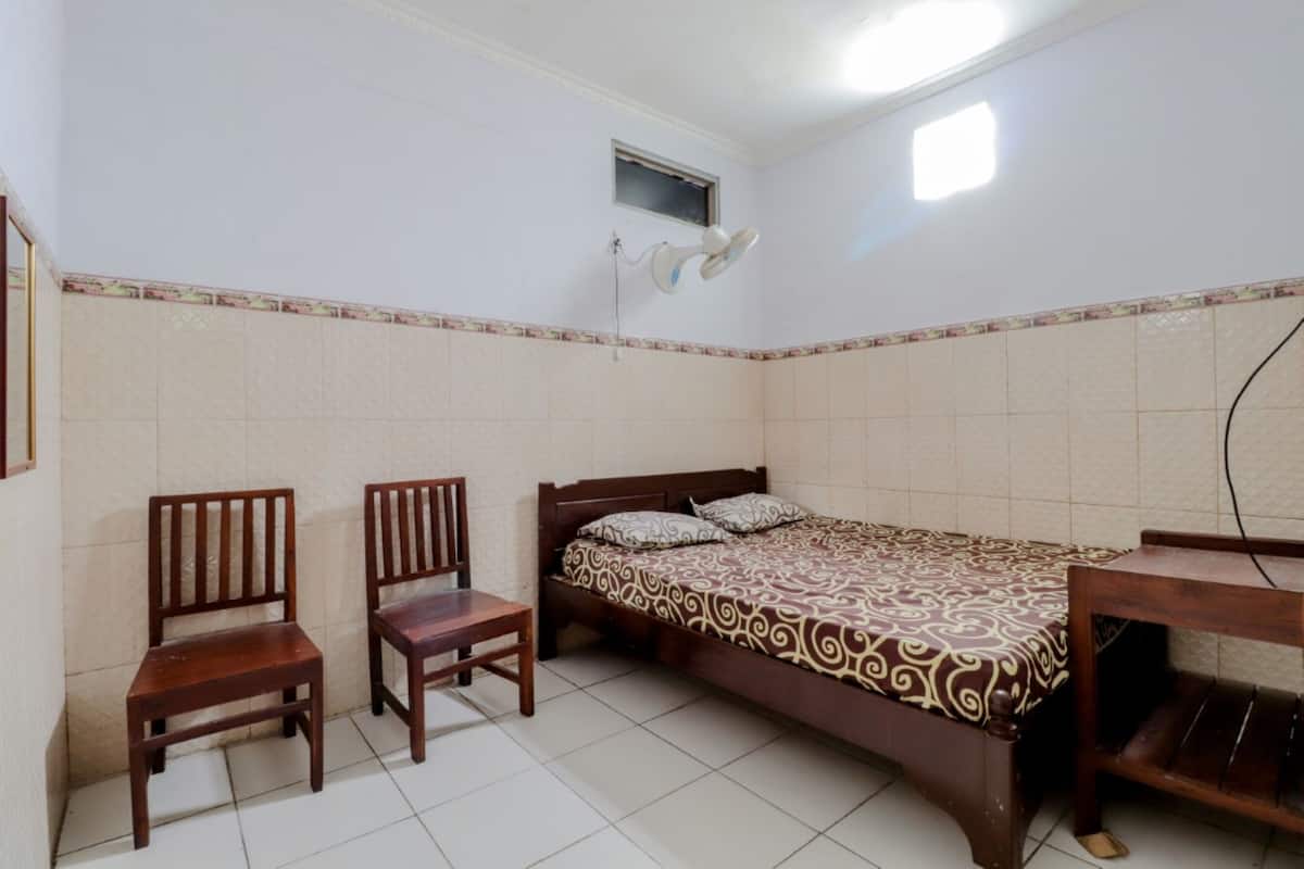 A small, clean room with a bed, two wooden chairs, a desk, and a wall-mounted fan.