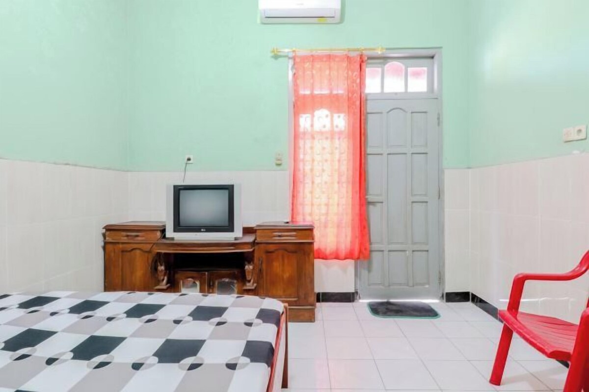 Una habitación con una cama, un escritorio con televisor, una silla roja y una puerta con cortina.