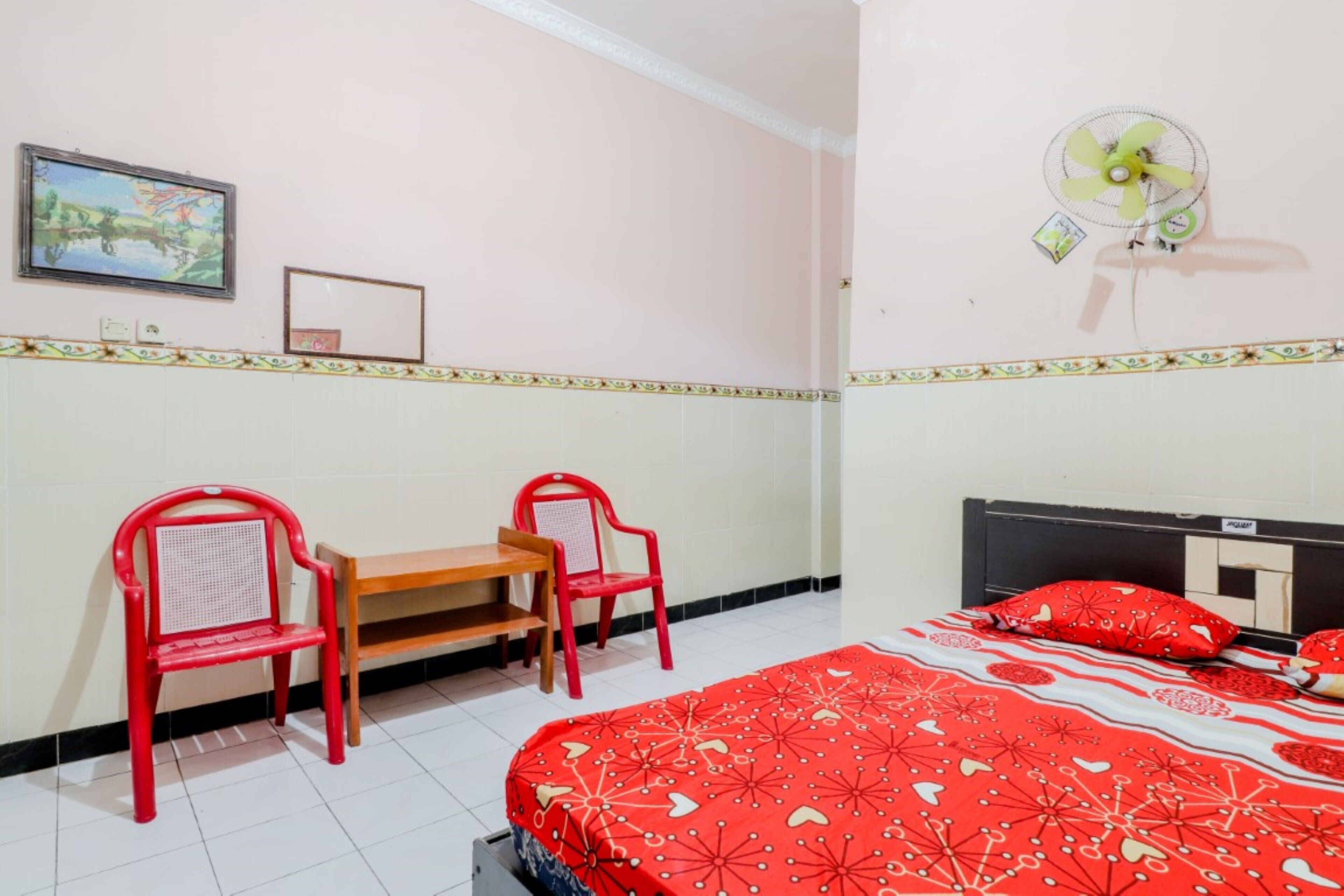 Une pièce comprenant un lit, deux chaises rouges, une petite table, un ventilateur fixé au mur et un tableau encadré.