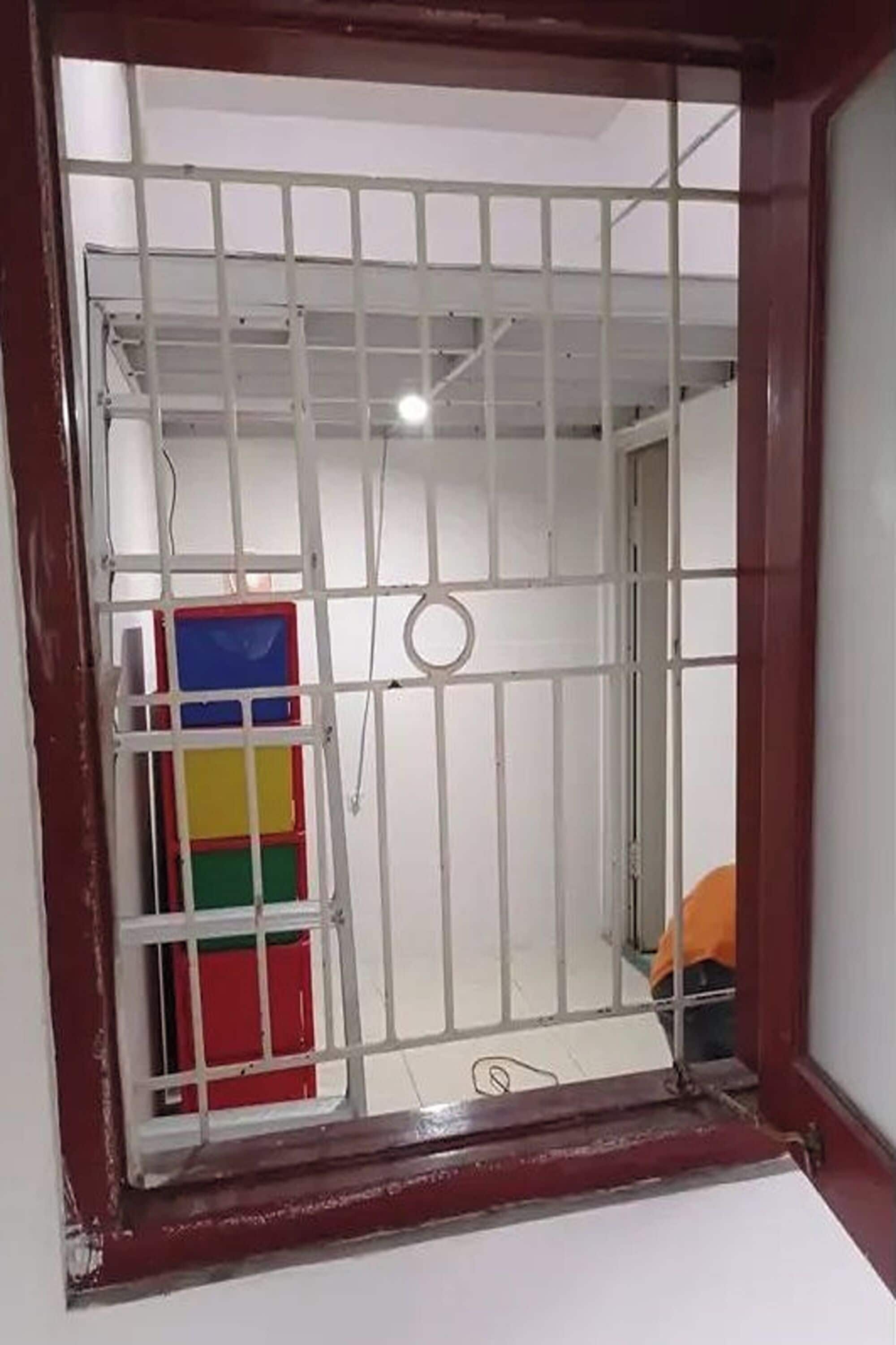 Quarto com janela com grades, cama com cobertor laranja e um móvel de armazenamento colorido.