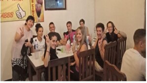 Un groupe de personnes est assis autour d’une table dans une pièce dont le mur est jaune et où est affichée une affiche publicitaire pour une boisson.