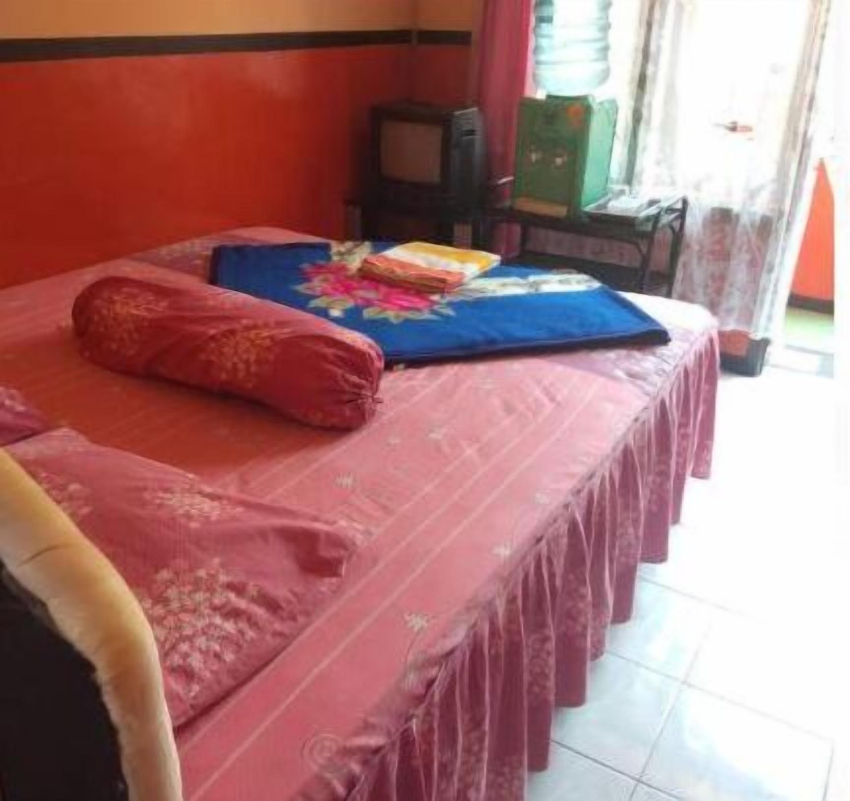 Une chambre avec un lit recouvert d’une couverture rose, d’une couverture bleue et d’un oreiller rouge. On y trouve une petite table sur laquelle sont posés une boîte verte et un objet rouge.