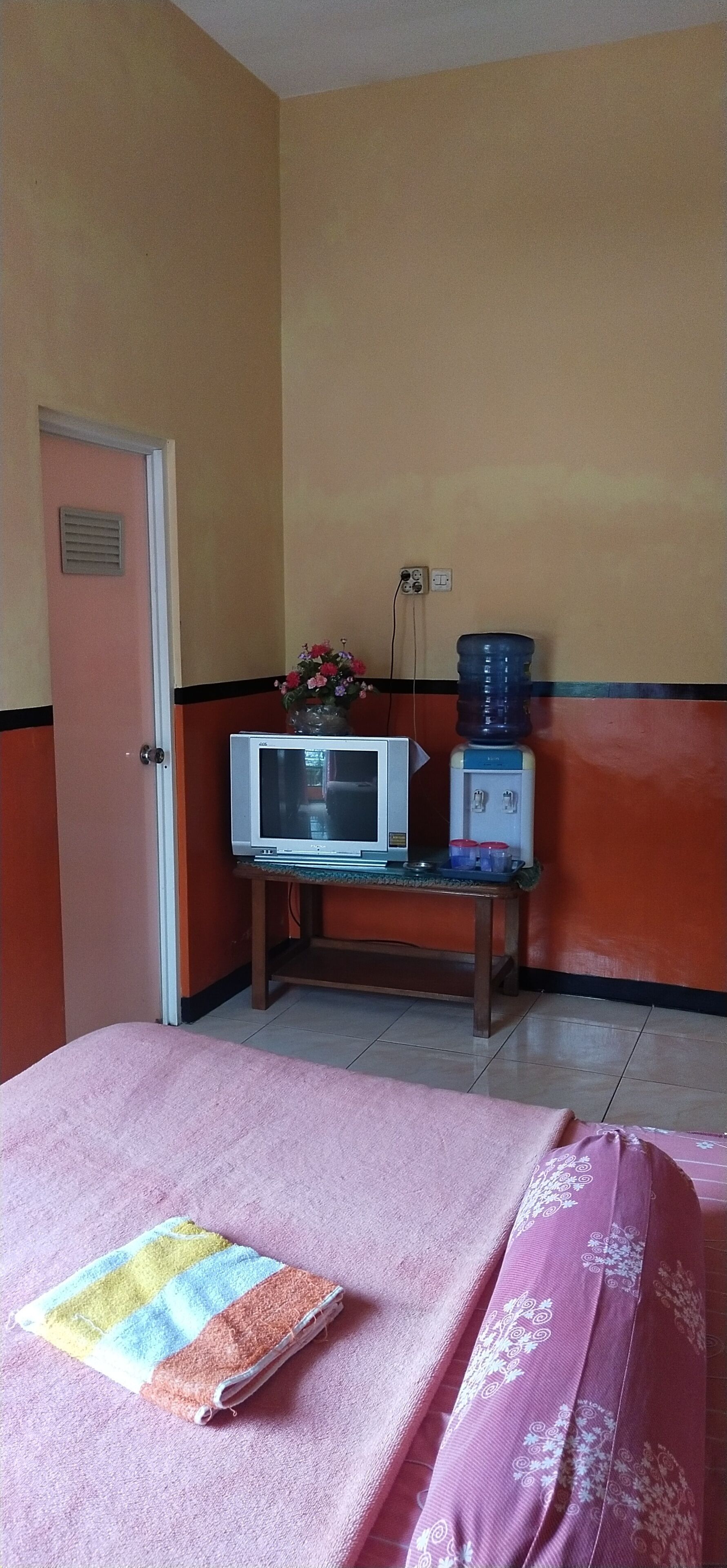 Quarto de hotel com cama, televisão em suporte, dispensador de água e uma porta.