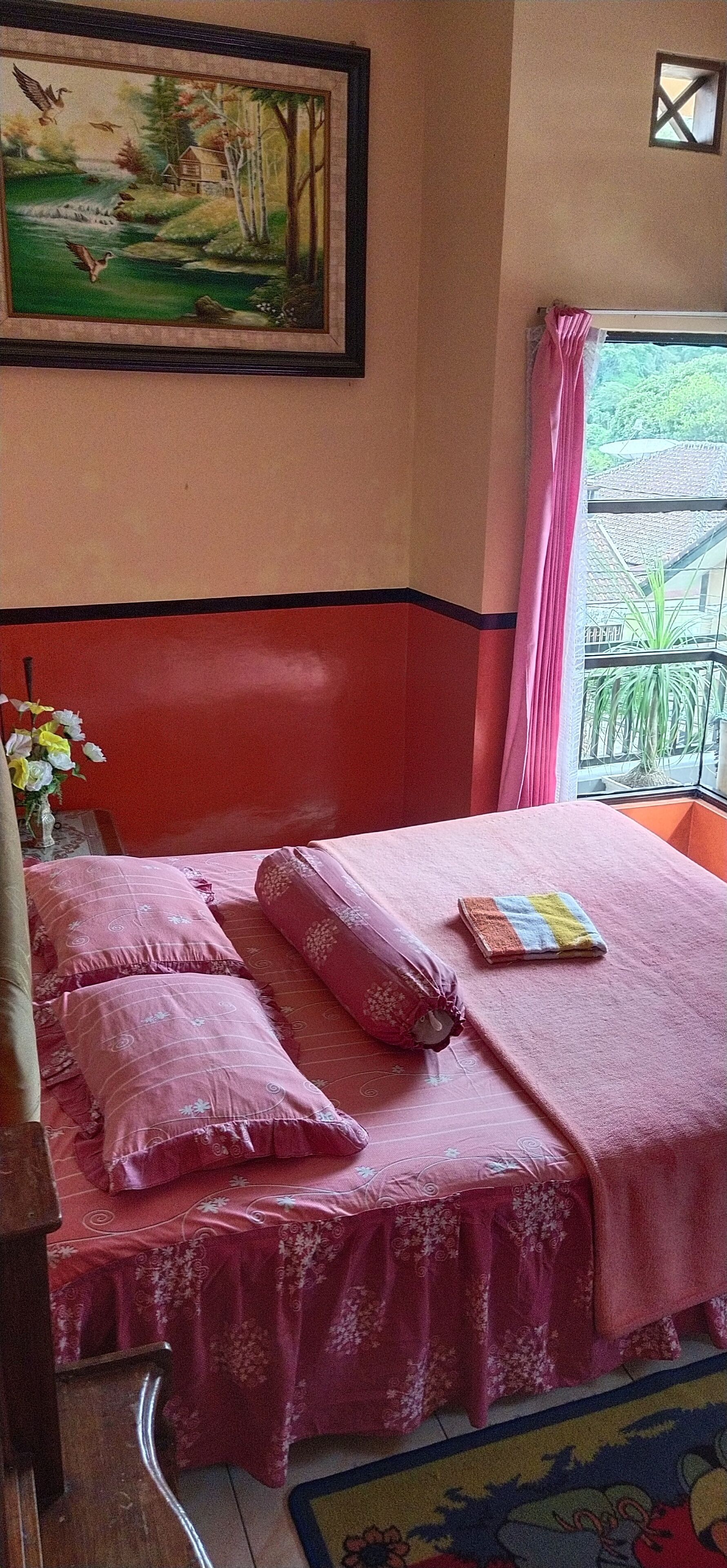 A bedroom with a bed covered in pink bedding, a painting of a landscape on the wall, and a window with a curtain.