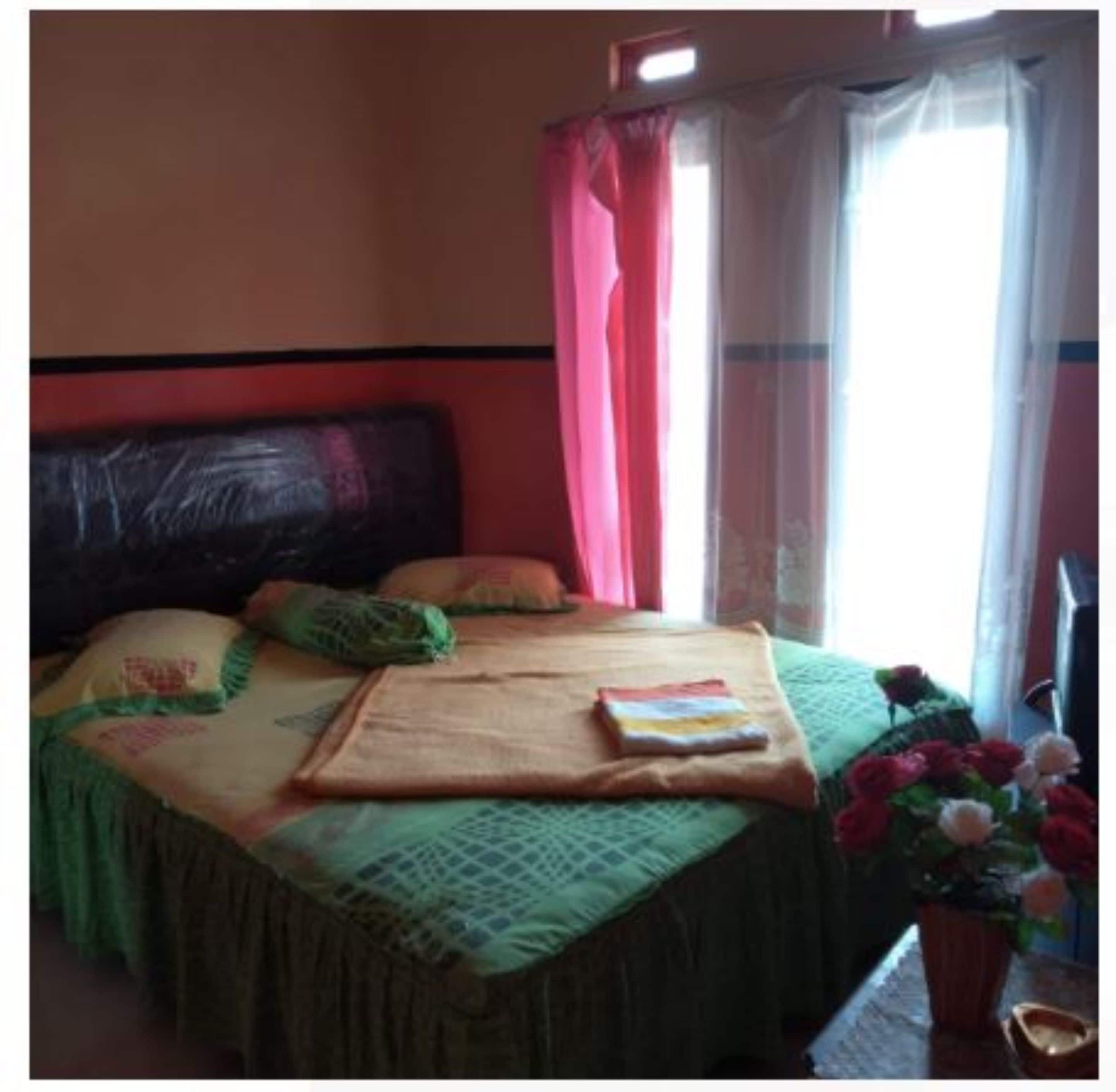 Ein Zimmer mit einem Bett, einem fensterbelaubten Fenster, einem kleinen Tisch mit einem Buch und einer Topfpflanze.