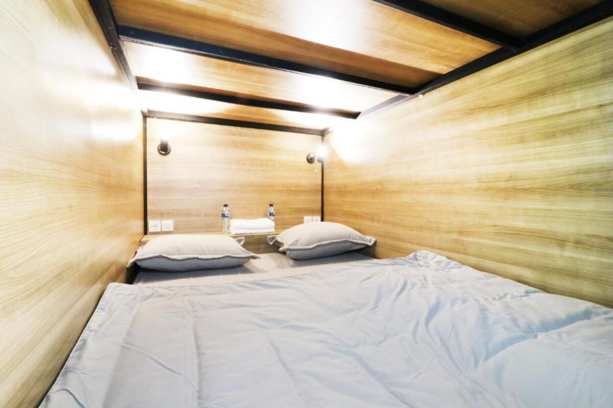 A compact, wooden-paneled room with a single bed, two pillows, a nightstand, and a wall-mounted light fixture.