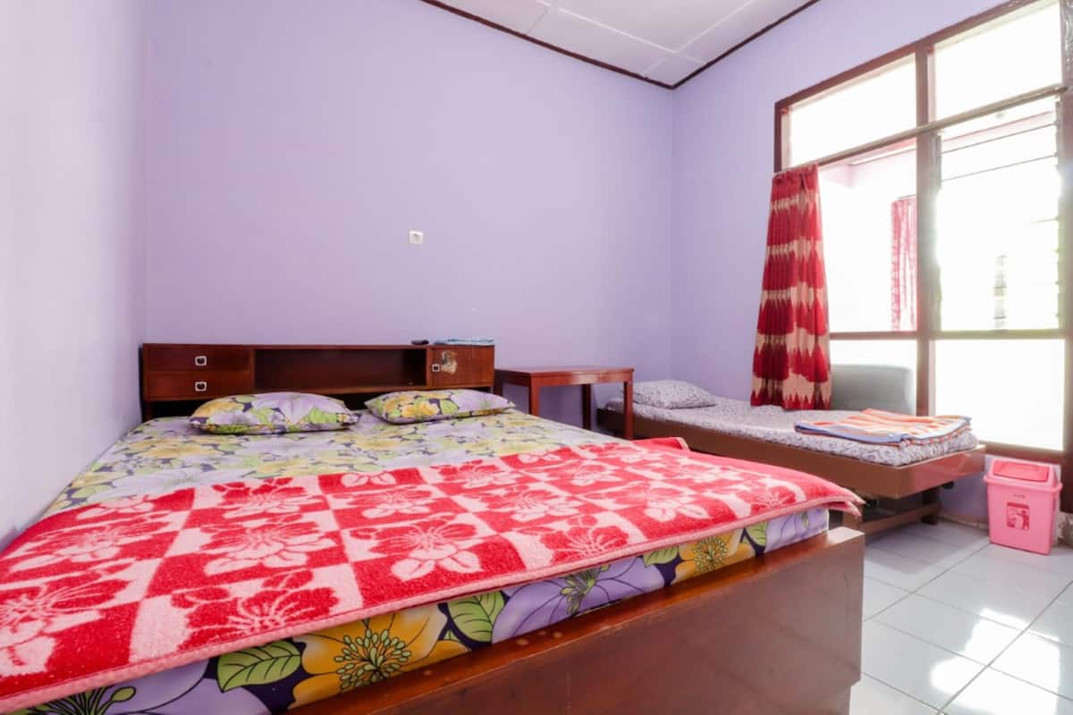 A bedroom with a bed, a small table, a window with curtains, and a trash bin.