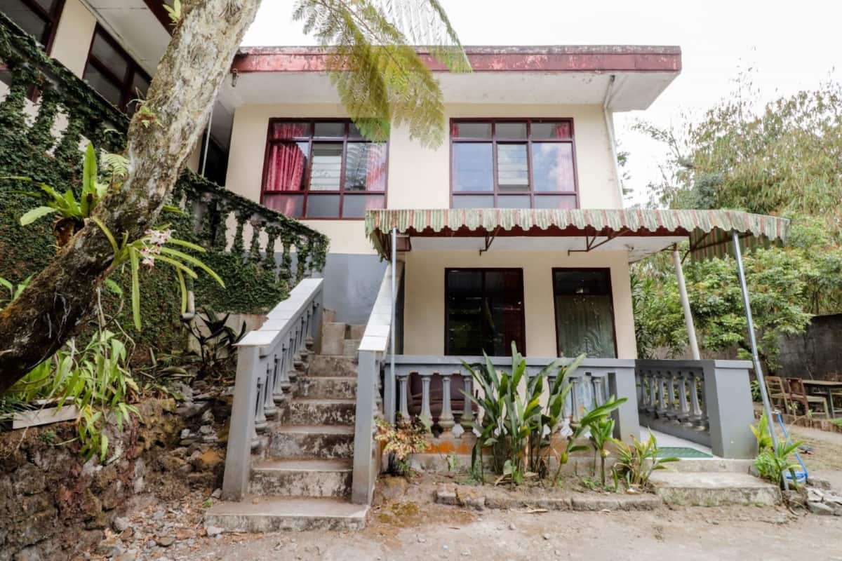 A two-story house with a staircase leading to the entrance, surrounded by greenery and a covered porch.