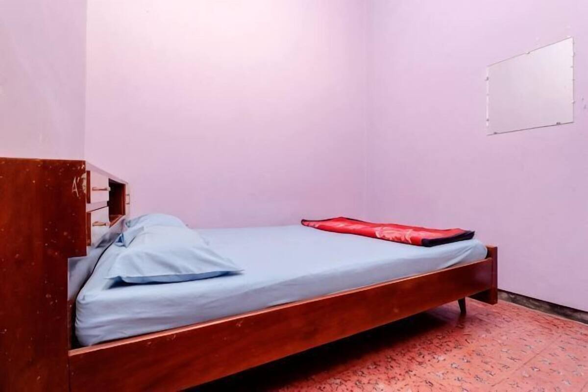 A single bed with a wooden frame, a blue mattress, and a red blanket in a room with pink walls and a patterned carpet.