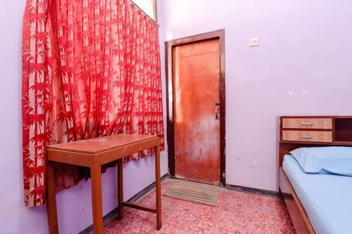 A room with a wooden desk, a bed with a blue bedspread, a red patterned curtain, and a wooden door.