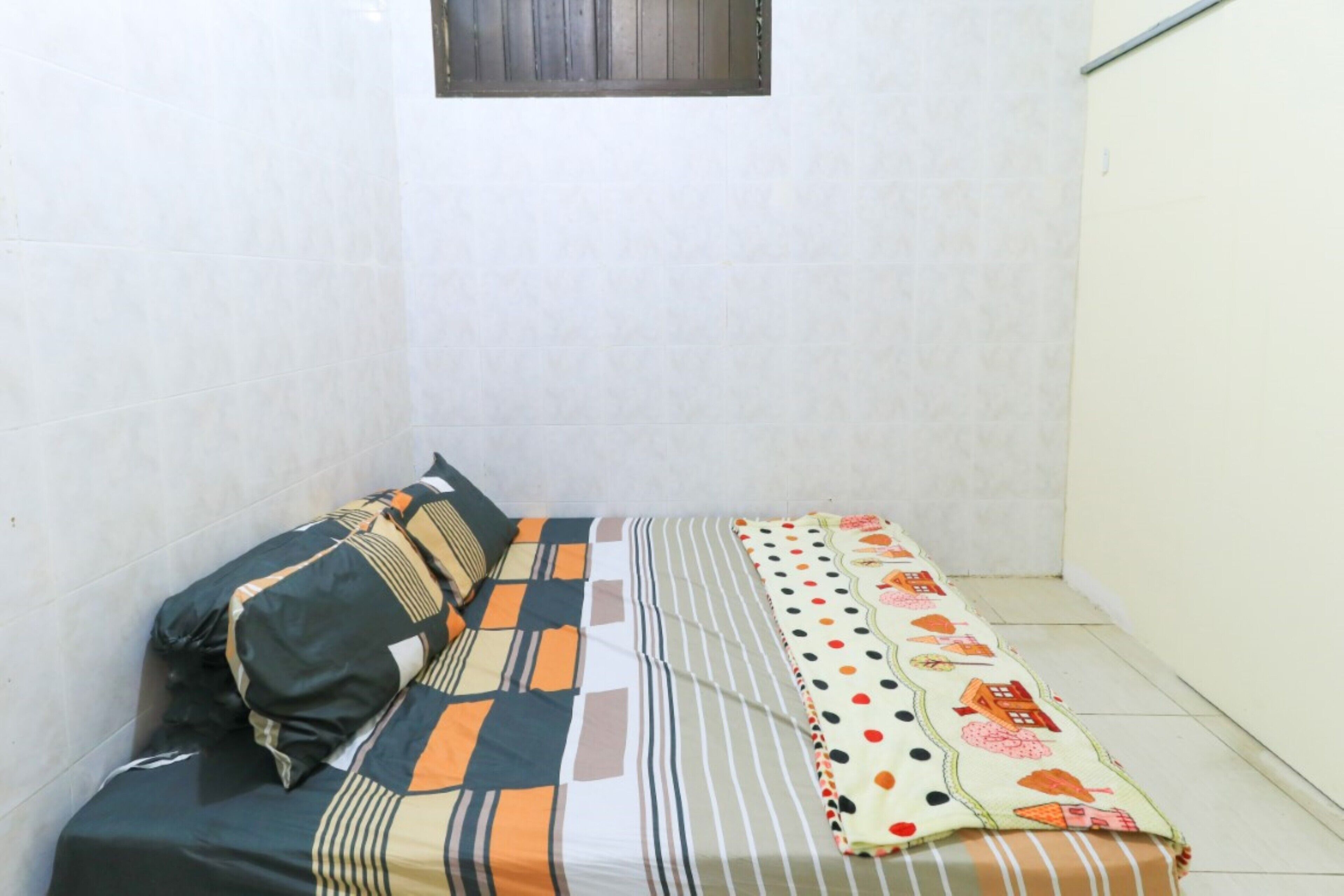 A small, narrow room with a bed against the left wall, a window above the bed, and a plain white wall on the right.