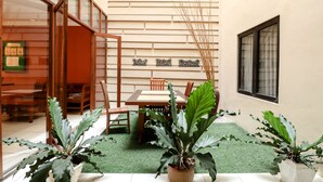 Uma área de refeições com mesa e cadeiras de madeira, cercada por plantas em vasos e um piso de grama sintética verde.