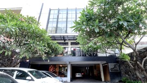 Um edifício moderno com fachada de vidro, entrada coberta e estacionamento com vários carros.