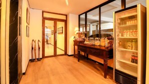 Um corredor com piso de madeira, porta de vidro que dá para outro quarto, uma escrivaninha de madeira com diversos objetos, e uma geladeira abastecida com bebidas.