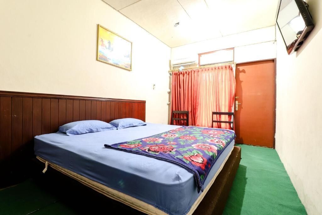 Un letto singolo con una coperta a fiori, una testiera in legno, un tappeto verde, un piccolo tavolino e una porta con tenda.