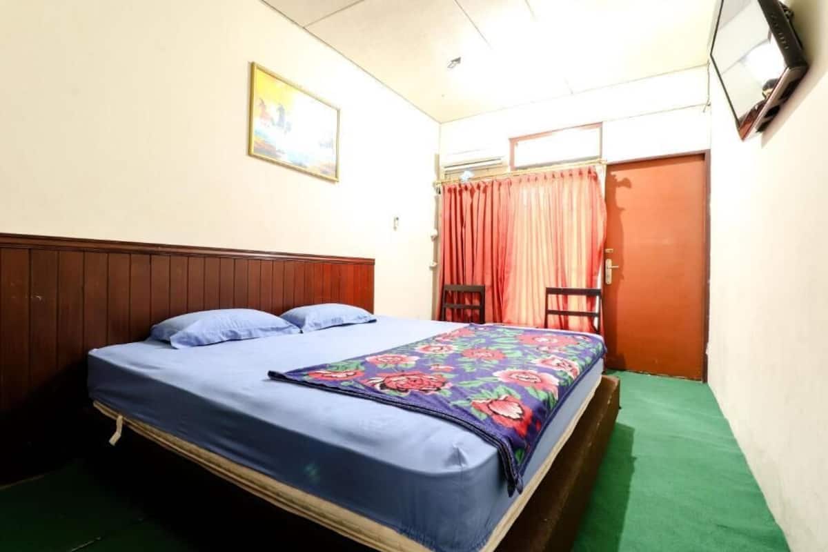 A single bed with a floral bedspread, a wooden headboard, a green carpet, a small table, and a door with a curtain.