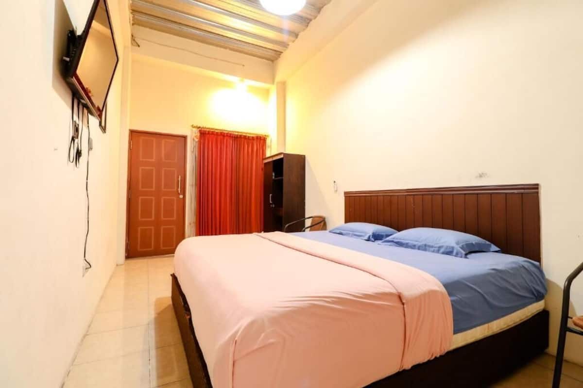 Un dormitorio con una cama, un televisor montado en la pared, una puerta y una cortina roja.