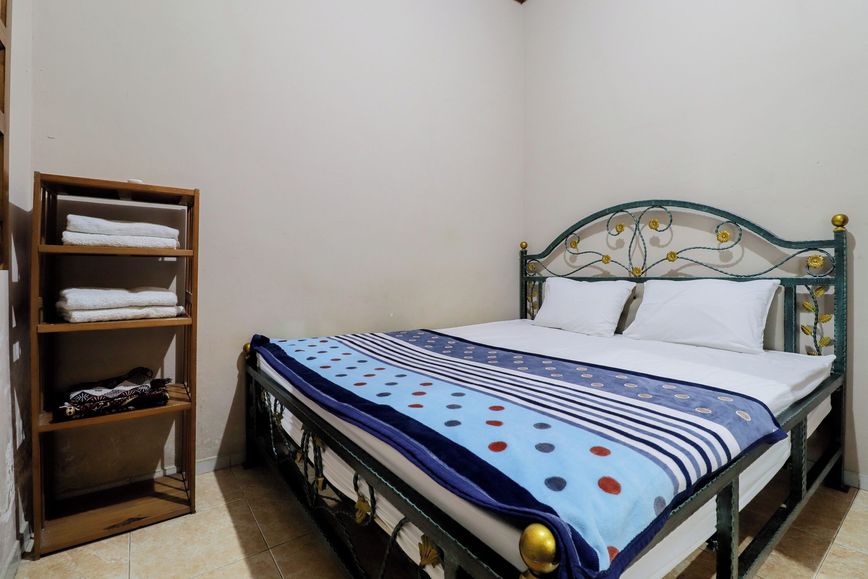 A single bed with a metal frame and a blue and white polka dot bedspread, a wooden shelf unit with folded towels, and a tiled floor.