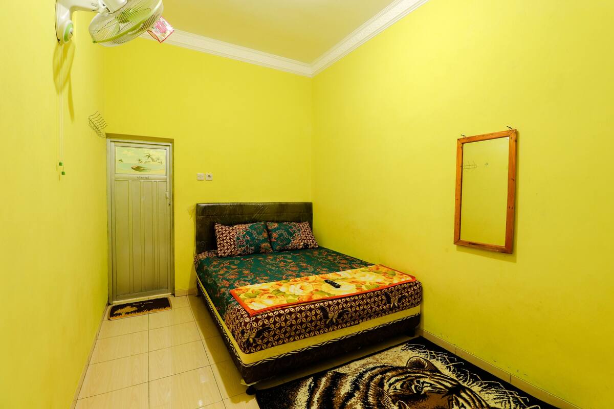 A small, brightly colored room with a bed, a door, a mirror, and a fan.