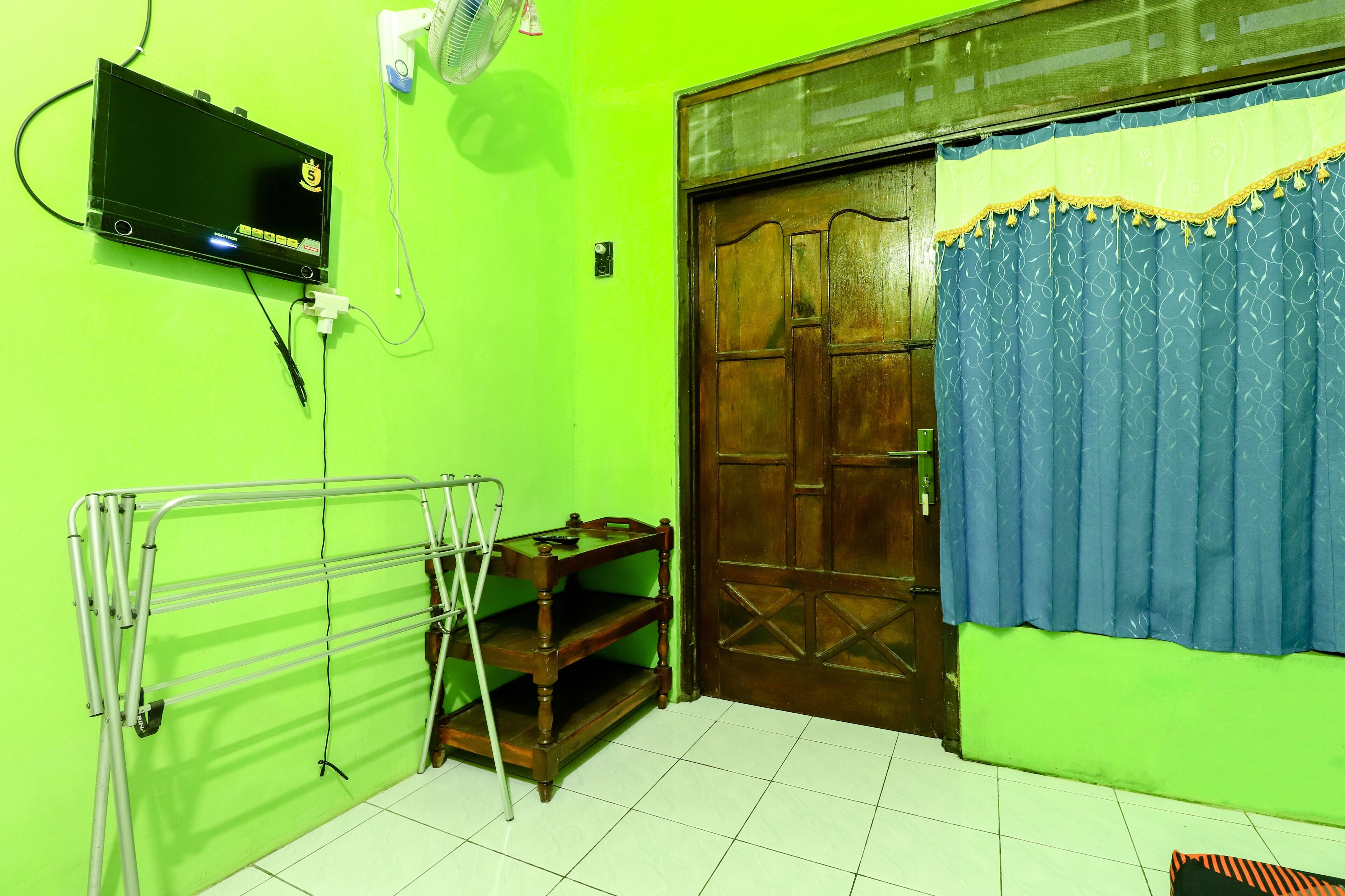 A room with a green wall, a wooden door with a blue curtain, a TV mounted on the wall, and a metal drying rack.