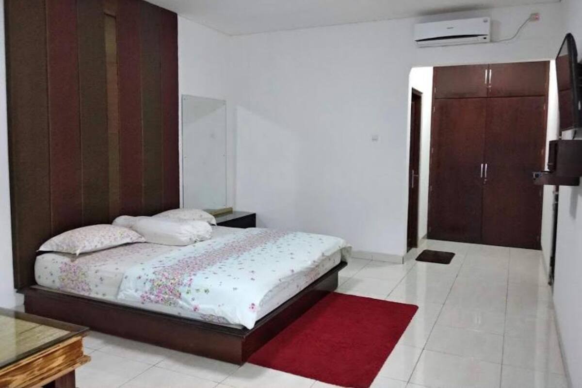 A bedroom with a bed, a red rug, a wardrobe, and an air conditioning unit.