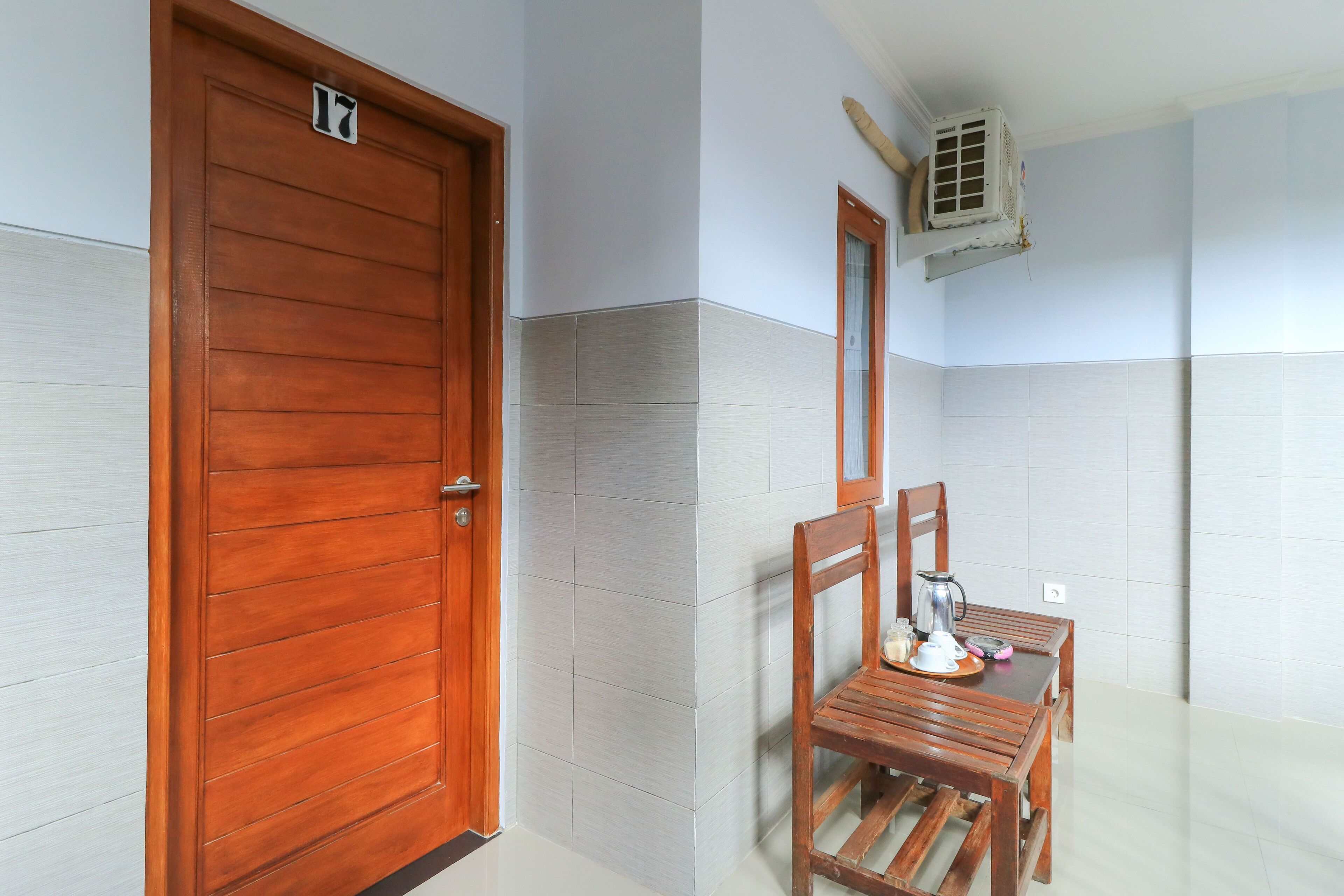 A hotel room with a wooden door, a tiled wall, a small table with a chair, and an air conditioning unit mounted on the wall.
