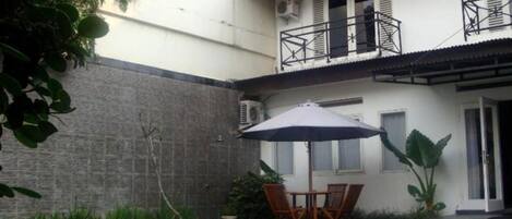 Una piscina con mesa y sillas bajo una sombrilla, rodeada de un jardín y un edificio con tejado de azulejos.