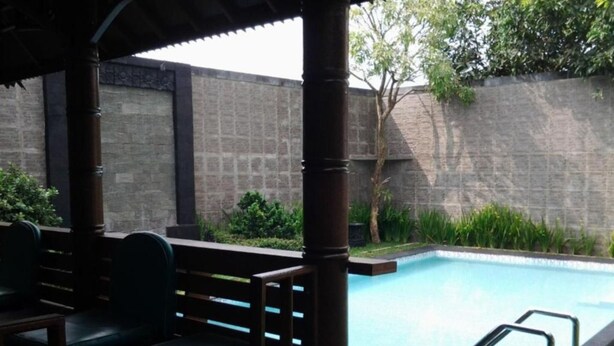 A pool area with a wooden deck, seating, and a stone wall in the background. - Rumah Anda Guest House (Bandung)