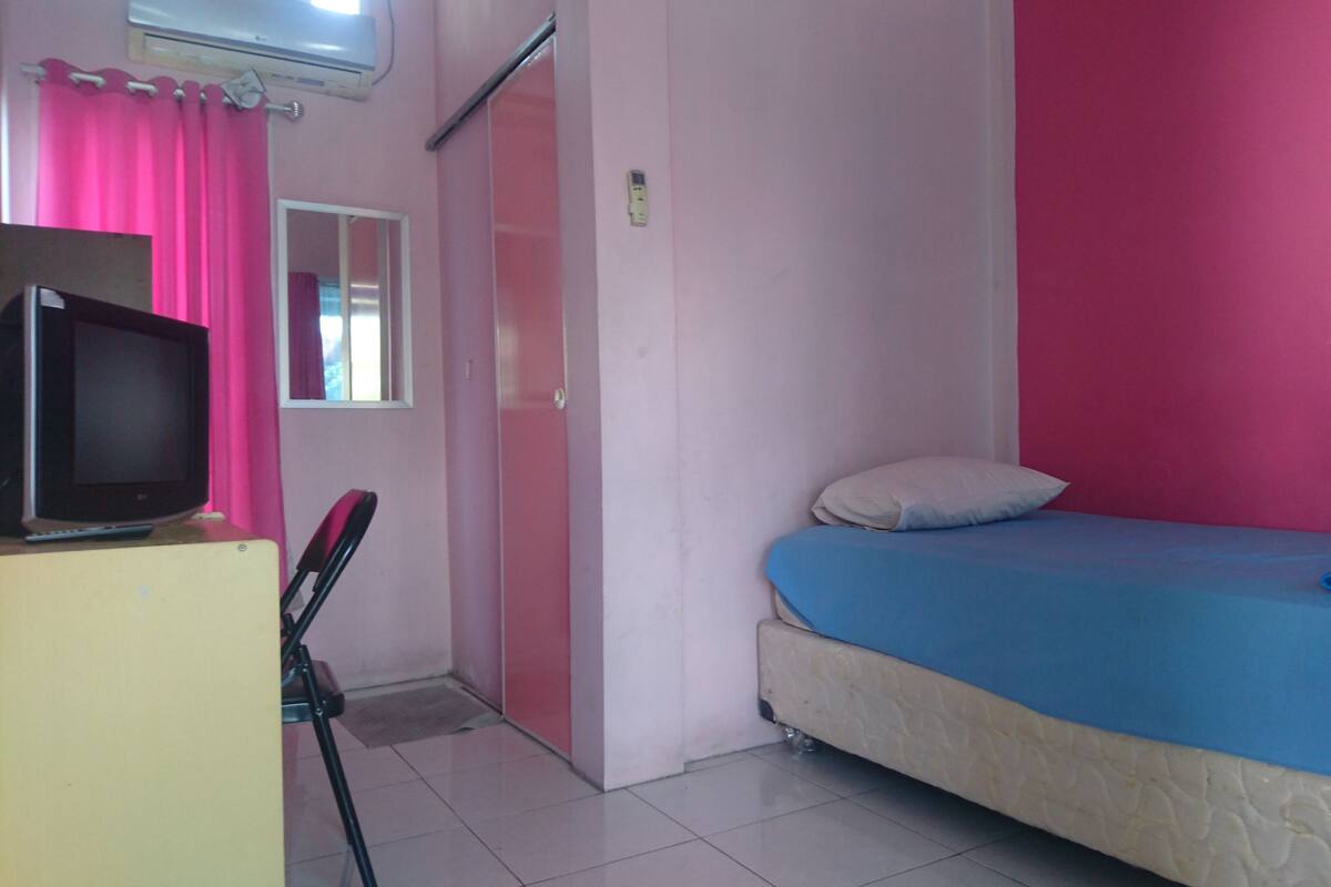 A room with a bed, a television on a desk, a window with pink curtains, and a door.