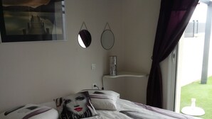 Un lit bien fait, recouvert d’une couvre-lit à motifs blancs et gris, avec deux oreillers et un coussin décoratif orné d’un motif représentant un visage. On peut voir deux miroirs fixés au mur et une petite étagère sur laquelle se trouve un récipient cylindrique. Une fenêtre avec des rideaux foncés est visible, et un tableau est accroché au mur.