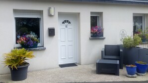 Une maison avec une porte blanche, trois fenêtres et un petit porche aménagé avec du mobilier d’extérieur.