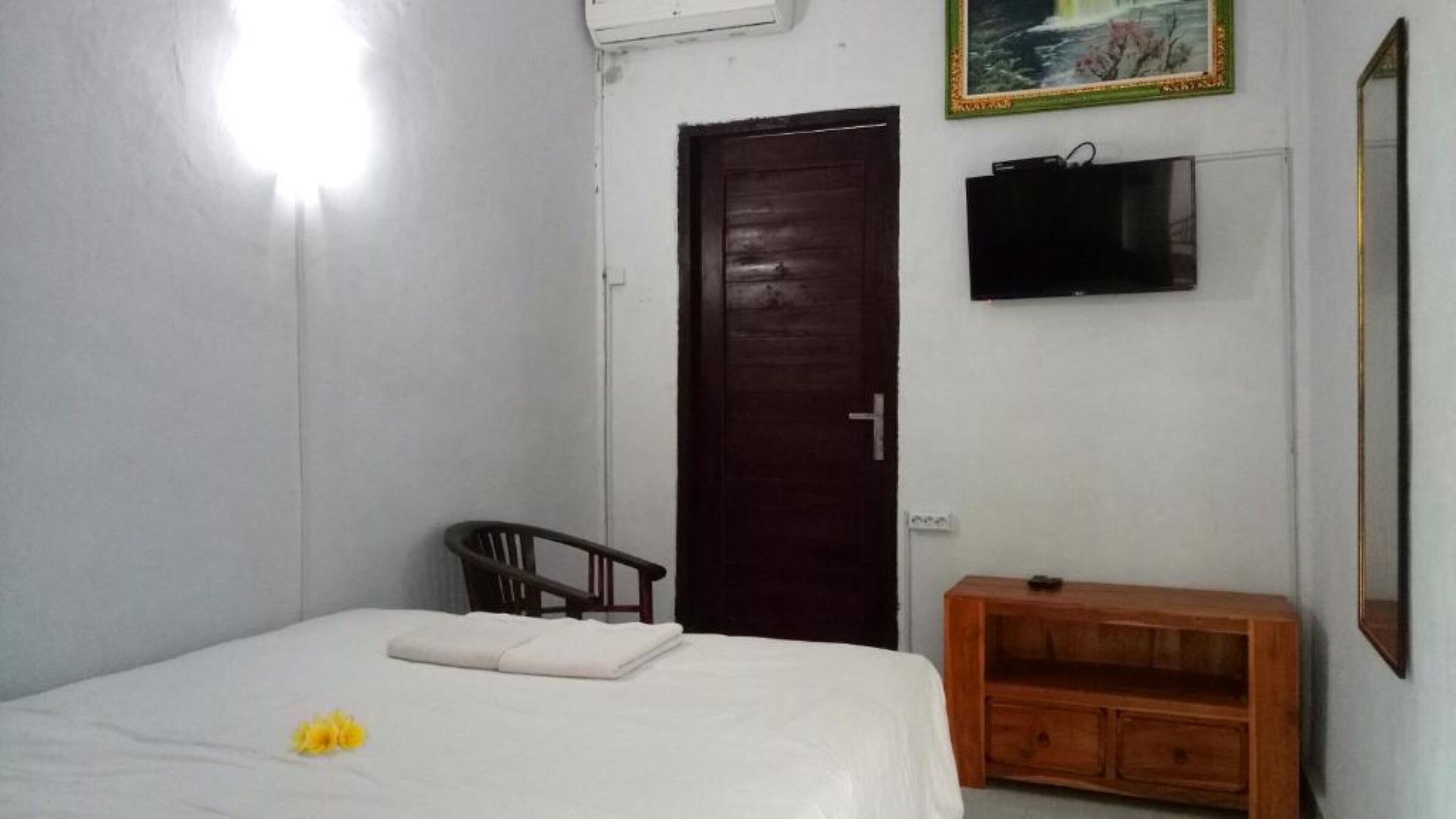 Un dormitorio con cama, mesita de noche de madera, silla, puerta, televisor y un cuadro enmarcado en la pared.