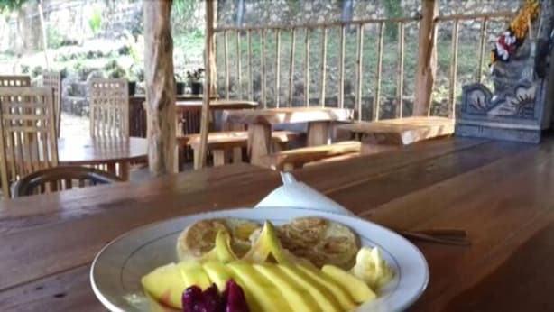 Een houten tafel met een bord eten, inclusief gesneden fruit en een afgedekte schaal, in een rustieke buitenomgeving met houten meubels en een leuning.
