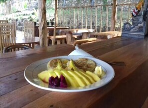 A wooden table with a plate of food, including sliced fruits and a covered dish, in a rustic outdoor setting with wooden furniture and a railing. - Broken Beach Villa (Penida Island)