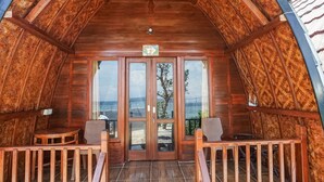 An interior view of a wooden cabin with a balcony, wooden flooring, and a view of the beach through glass doors. - Road Beach Villa (Penida Island)
