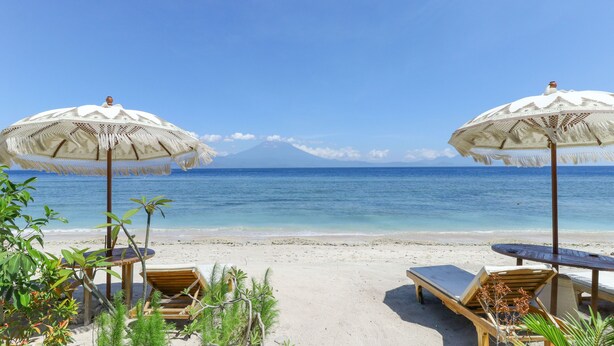 A beach with two large umbrellas, wooden lounge chairs, and a view of the ocean. - Road Beach Villa (Penida Island)