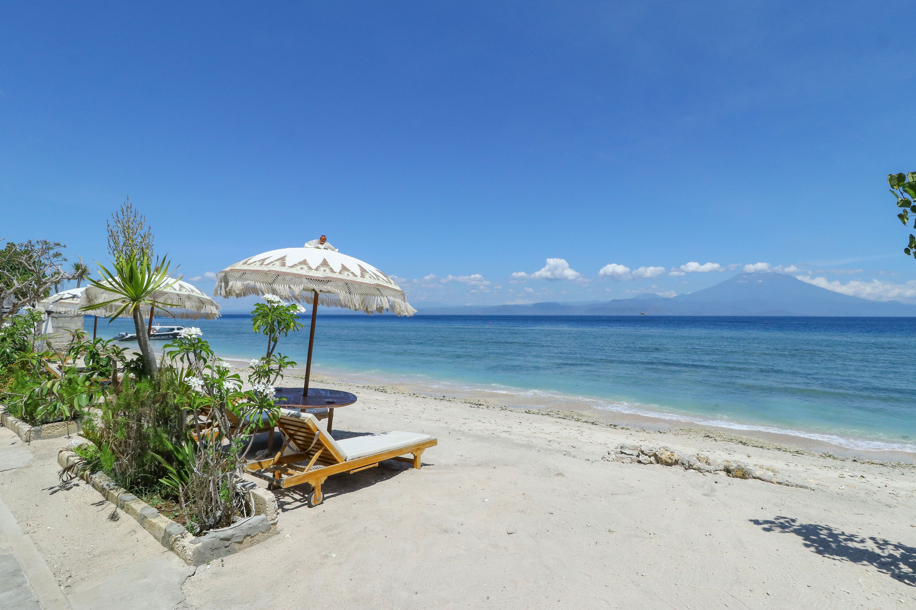A beach with a wooden lounge chair and a thatched umbrella, a clear blue sky, and a mountain in the distance.
