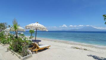 A beach with a wooden lounge chair and a thatched umbrella, a clear blue sky, and a mountain in the distance.