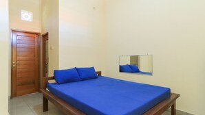 Une chambre avec un lit en bois, un matelas bleu, une tête de lit en bois, un petit miroir accroché au mur et une porte surmontée d’un luminaire.