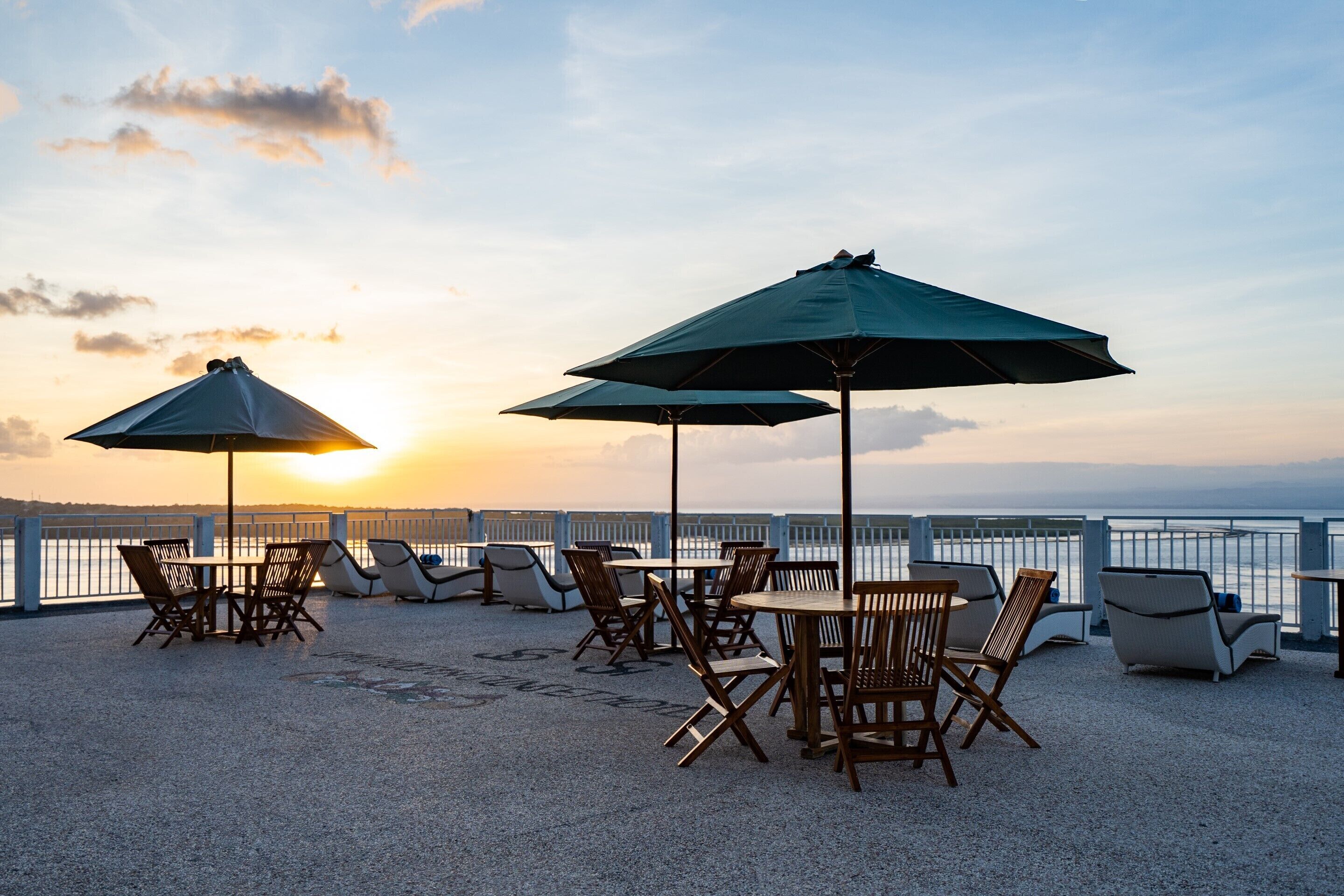 A terrace with tables and chairs, umbrellas, and a view of the sea at sunset.