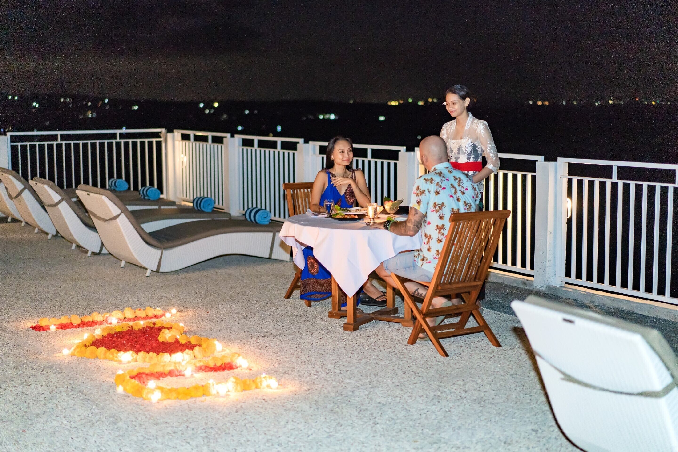 A dining area with a table set for two, surrounded by lounge chairs, and a decorative arrangement of candles and flowers on the floor.