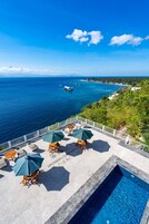 A pool area with lounge chairs and umbrellas, overlooking a coastal view with boats and greenery.