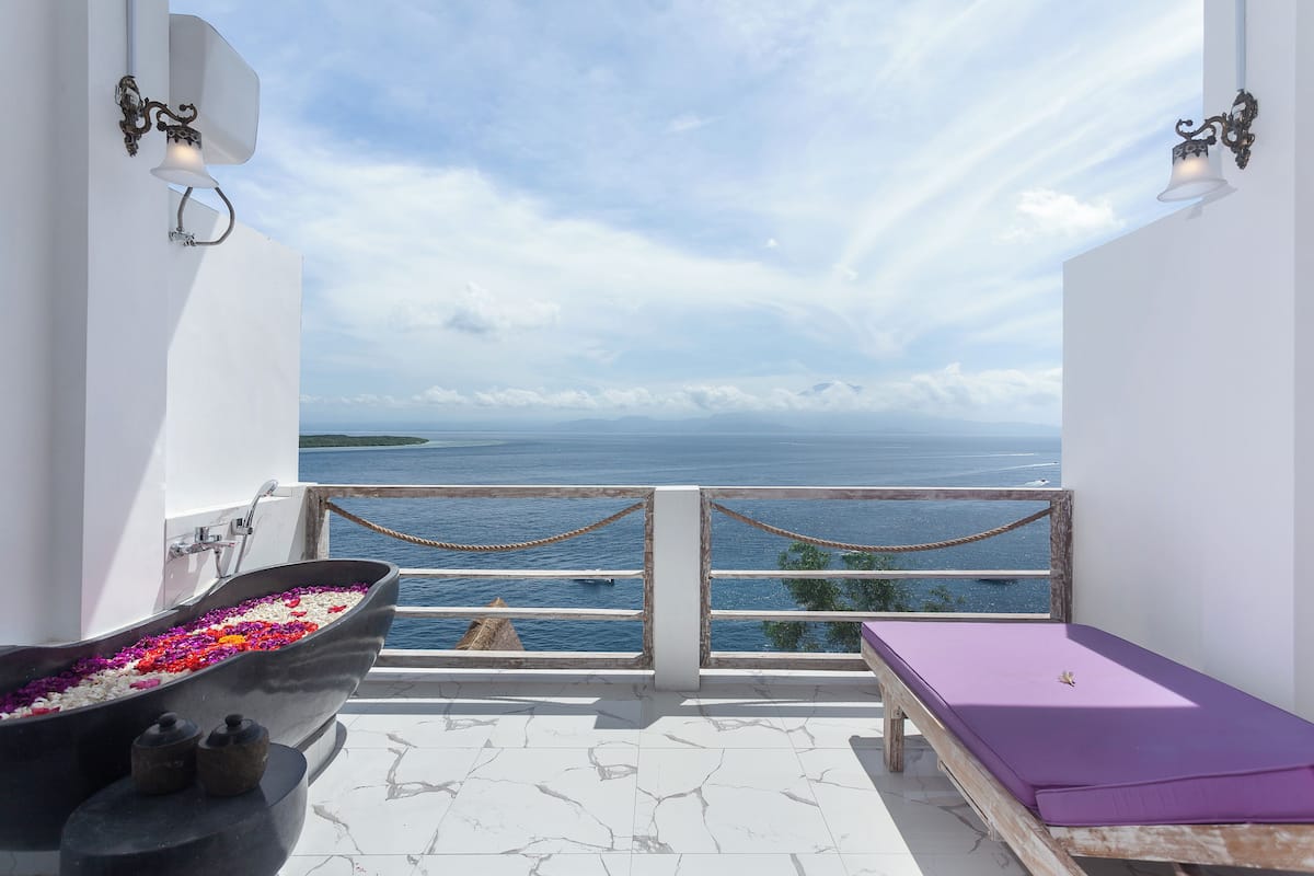 Un balcón con vistas al mar, una bañera llena de flores y un banco de madera con un cojín morado.