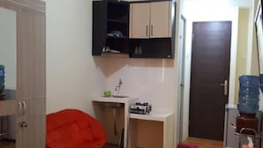 Una habitación compacta con una silla roja, un escritorio pequeño con una computadora, un dispensador de agua y una puerta.