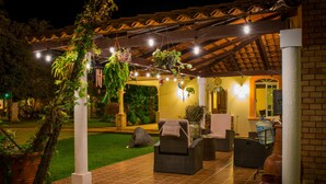 Uma área de estar externa com piso de azulejos, móveis de vime, plantas penduradas e uma parede de tijolos.