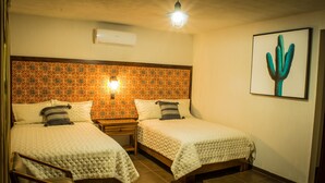 Quarto de hotel com duas camas, uma mesinha de cabeceira, uma cadeira, ar-condicionado fixado na parede e uma obra de arte com cactos.